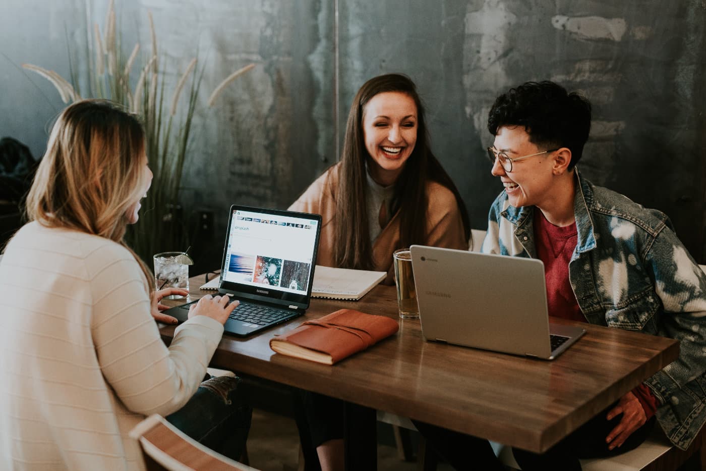 Three women sit at a desk with their laptops. They are chatting and laughing.